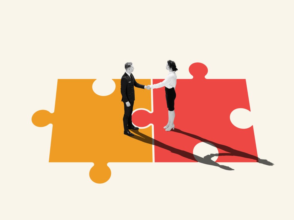 Two business people standing on different but complementary puzzle pieces and shaking hands - representing partnership and successful business acquisition.